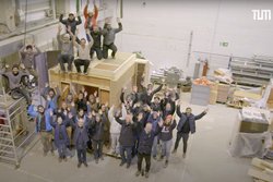 Screenshot eines Videos. Eine Menschengruppe von ca. 20 Personen steht in einer Werkstatt. Sie schauen in die Kamera und halten die Arme in die Luft. 