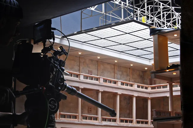 The silhouette of a professional film camera can be seen in the left foreground. Behind it is a bright film studio with skylights.