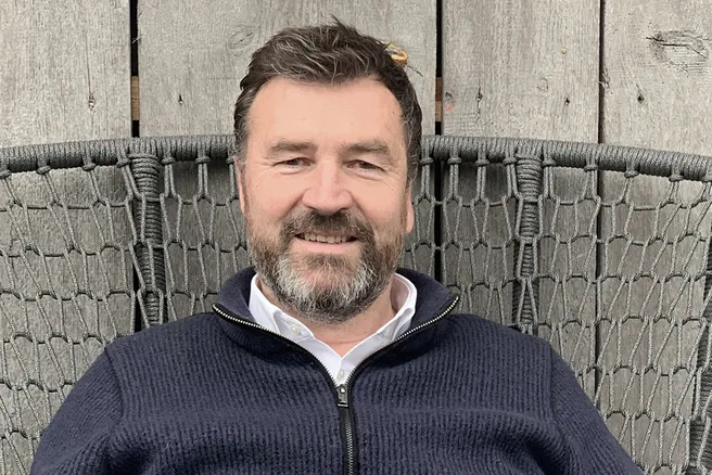 Portraitfotografie von Florian Nagler der ein weißes Hemd unter einer dunkelblauen Strickjacke trägt und vor einem holzgetäfelten Hintergrund sitzt.