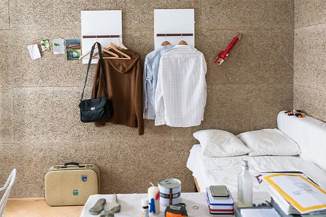[Translate to en:] Holzwerkstoffplatten mit Kleiderhaken und Kleidern, rechts Bett, im Vordergrund Bücher und private Gegenstände
