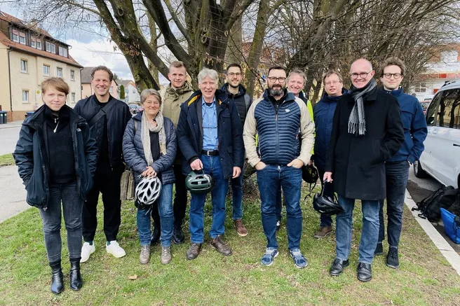 Gruppenfoto des Projektteams. Außen aufgenommen, sie stehen auf einer kleinen Grünfläche, urbaner Raum im Hintergrund zu erkennen.